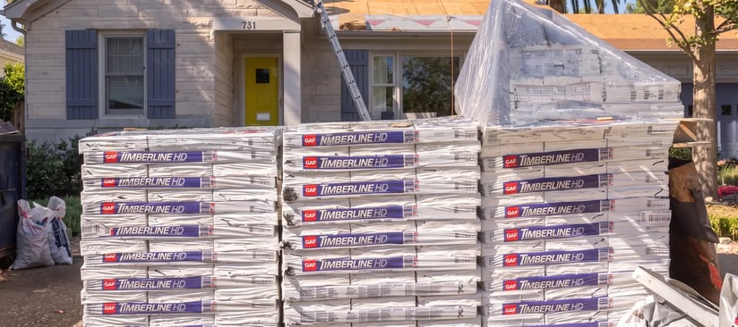 Stacked asphalt shingle bundles staged in a driveway with the roof deck exposed during a re-roof