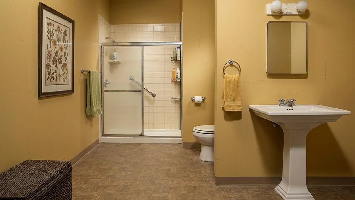 Dated framed shower door, gold walls and worn vinyl flooring before a bathroom remodel
