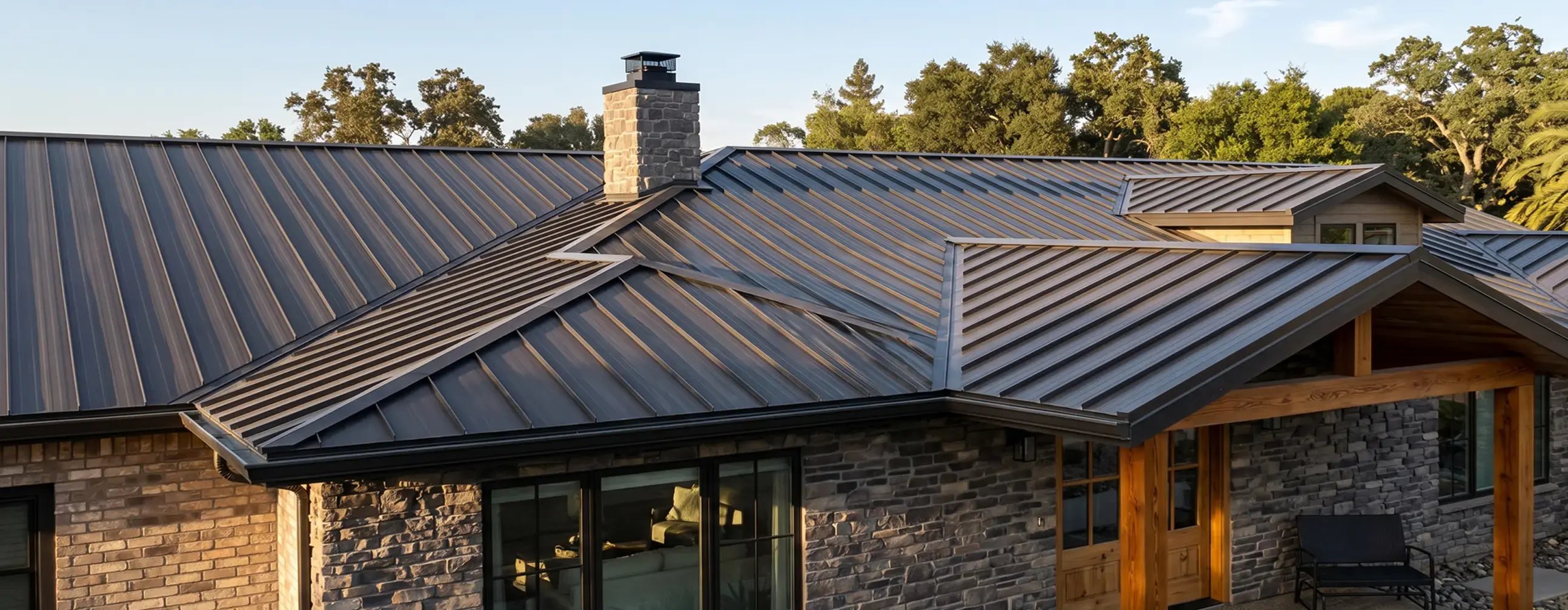 Dark bronze standing seam metal roof on a stone and brick single-story home at dusk
