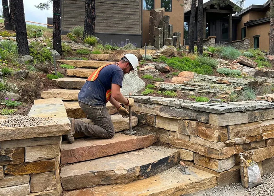 Worker setting natural stone steps and a stacked-stone retaining wall on a landscaped hillside under construction
