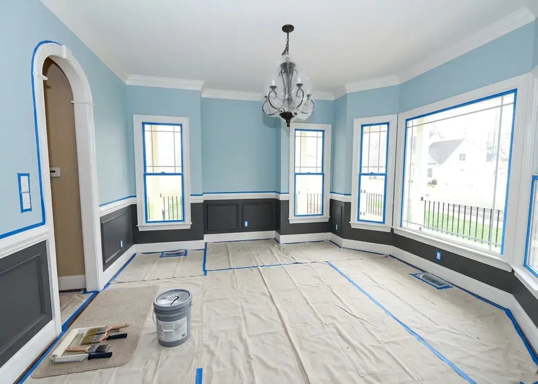 Two-tone blue and gray dining room walls with taped trim and floor cloths during interior painting — Hybrid Renovations SEO Reasoning: Describes the two-tone walls and masking that signal active interior painting, distinct from the exterior card. Branded. Uncertainty: None. Batch check: 3 of the 5 are branded (stucco, exterior paint, interior paint) and 2 are not (roofing, insulation), and every alt text is worded differently. I used .webp in the file names to match your uploaded files; if your CMS serves these as .jpg, swap the extension and everything else stays the same.