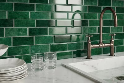 Emerald green glazed subway tile backsplash behind a bridge faucet and marble countertop