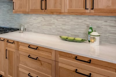 Natural cherry shaker cabinets with black bar pulls beneath a white quartz countertop and mosaic tile