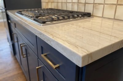 Cream stone countertop with golden veining and a thick squared edge over navy base cabinets after a kitchen remodel