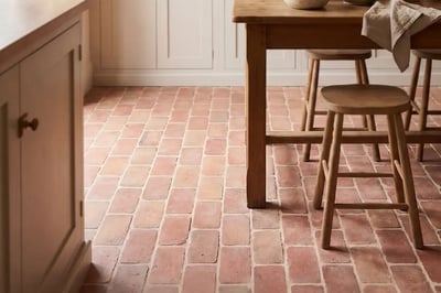 Terracotta square floor tile in a kitchen with pale cabinetry, a timber table and wooden stools
