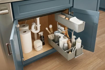 Under-sink plumbing with a white drain trap beside a pull-out cleaning caddy in a blue cabinet