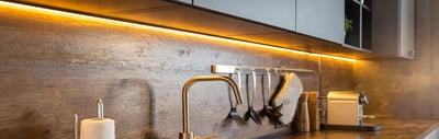 Warm LED strip lighting under kitchen wall cabinets washing across a wood plank backsplash
