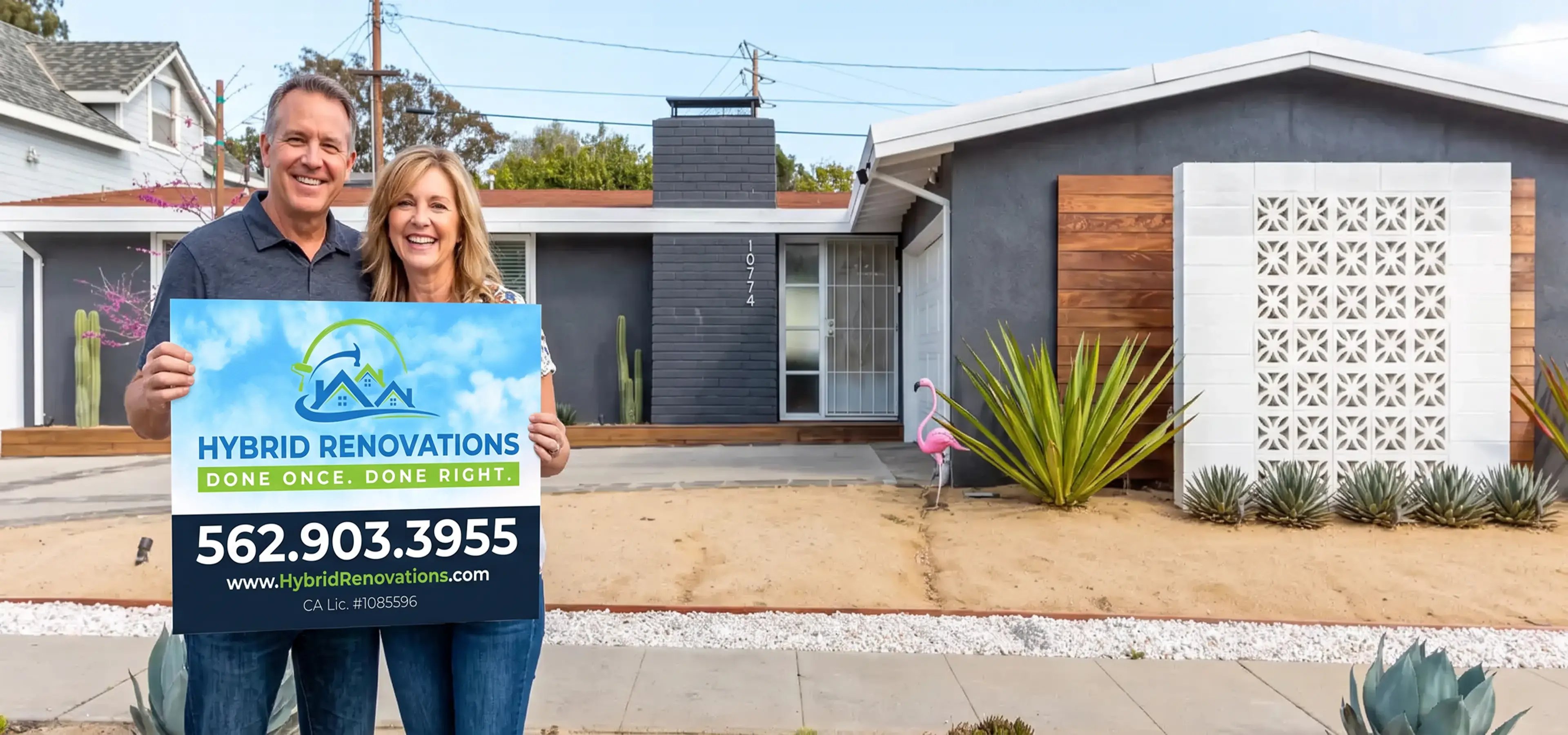 Smiling homeowners holding a Hybrid Renovations yard sign in front of their renovated mid-century home