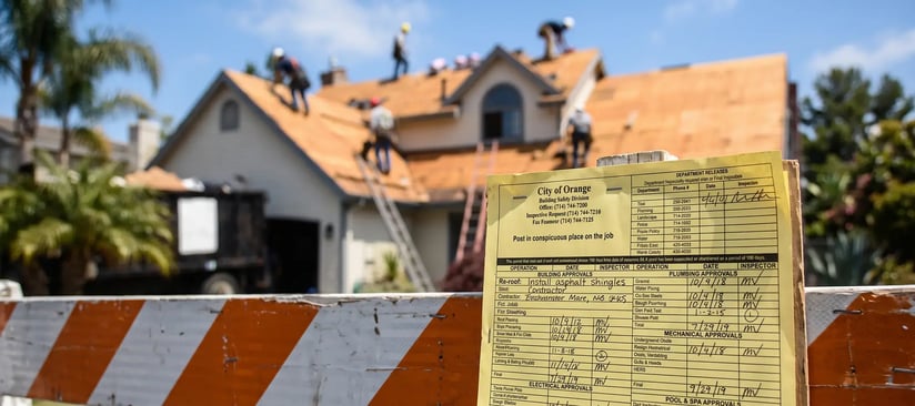 Building permit card posted at a re-roof job site with a crew installing sheathing on the house behind