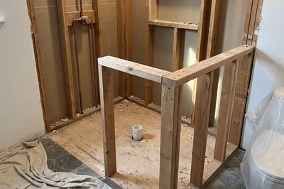 Shower demolished to bare studs with the knee wall framed and drain stub exposed in the subfloor