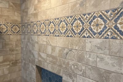 Travertine-look wall tile with a blue and gold decorative accent band after a shower rebuild